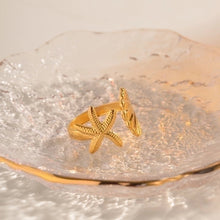 Load image into Gallery viewer, Gold starfish and seashell ring styled on a clear textured glass dish, capturing its radiant shine. The adjustable open ring design features 18K gold plating over stainless steel, waterproof and tarnish resistant for lasting wear. Inspired by the sea, this ring makes a chic coastal accessory, ideal as everyday jewellery or as a unique ocean themed gift for women.