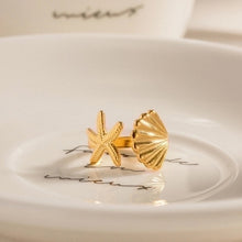 Load image into Gallery viewer, Isla Starfish Seashell Ring displayed on a white ceramic dish with cursive lettering, highlighting its detailed gold starfish and scallop shell design. The 18K gold plated stainless steel ring has an adjustable fit, making it versatile for everyday wear. Inspired by the ocean, this waterproof ring is a playful yet elegant piece of jewelry for women, ideal for gifting.