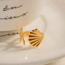 Load image into Gallery viewer, Starfish and seashell ring placed beside a white ceramic cup, showing the hammered scallop shell and textured starfish details. Crafted from 18K gold plated stainless steel, this adjustable ring combines durability with feminine ocean inspired style. A waterproof jewellery piece perfect for beach lovers, summer wear, or as a gift for women who love coastal jewellery.