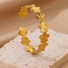 Load image into Gallery viewer, Gold flower cuff bracelet displayed standing on a beige stone surface. The adjustable floral bangle features hammered 18K gold plated petals with detailed textures, giving the bracelet a boho yet luxurious look. Designed from waterproof stainless steel, this open cuff bracelet makes an elegant jewellery gift for women and can be styled as a standalone statement or stacked with other bangles for a chic gold cuff stack.