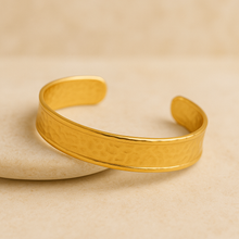 Load image into Gallery viewer, MJ Luxe Co Colette Gold Textured Cuff Bracelet photographed at an angle on a stone backdrop, highlighting its hammered finish and 18K gold plated stainless steel design. This open cuff bangle is adjustable, waterproof, tarnish resistant, hypoallergenic, and nickel free, making it an ideal minimalist gold bracelet for daily wear, layering, or gifting as a bridal or bridesmaid jewellery piece.