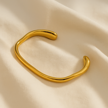 Load image into Gallery viewer, MJ Luxe Ava Gold Cuff Bracelet displayed on cream fabric, minimalist modern jewelry design with sculptural curved silhouette.