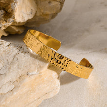 Load image into Gallery viewer, Close-up of the MJ Luxe Hammered Gold Cuff Bracelet resting on natural stone. The 18K gold plated stainless steel cuff features a hammered texture with a polished shine, photographed in soft natural light to highlight its sculptural detail. Waterproof, tarnish-resistant, minimalist adjustable bangle for everyday wear or layering with other bracelets.

