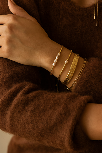 Elegant model wearing the MJ Luxe Hammered Gold Cuff Bracelet stacked with dainty gold and pearl bracelets, styled with a brown knit jumper for a luxe everyday look. The textured hammered cuff adds bold shine while blending seamlessly with minimalist, stackable jewellery. 