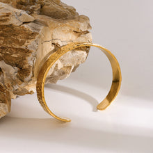 Load image into Gallery viewer, The MJ Luxe Hammered Gold Cuff Bracelet leaning against a natural rock with soft shadows and warm light. The hammered detailing adds artisanal charm while the polished 18K gold plated stainless steel ensures durability. Waterproof and tarnish resistant, this minimalist open cuff is the perfect layering bracelet or modern gold statement bangle.
