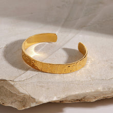 Load image into Gallery viewer, Flat side view of the MJ Luxe Hammered Gold Cuff Bracelet displayed on soft natural stone. The sleek hammered texture and high-polish gold plating reflect the light beautifully, showing the bold yet minimal silhouette. Adjustable, stackable, waterproof 18K gold cuff designed for timeless everyday jewelley and luxury minimalist styling.
