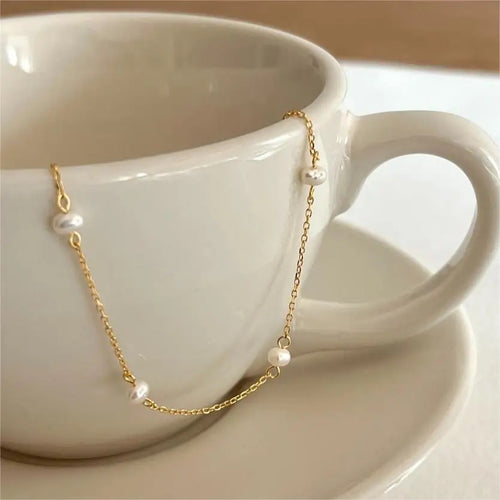A gold chain bracelet with freshwater white pearls is displayed draped over the edge of a white coffee cup.