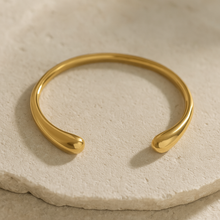 Load image into Gallery viewer, MJ Luxe Ava Gold Cuff Bracelet displayed on beige stone surface, minimalist sculptural open bangle with polished golden finish.

