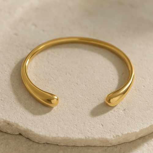 MJ Luxe Ava Gold Cuff Bracelet displayed on beige stone surface, minimalist sculptural open bangle with polished golden finish.