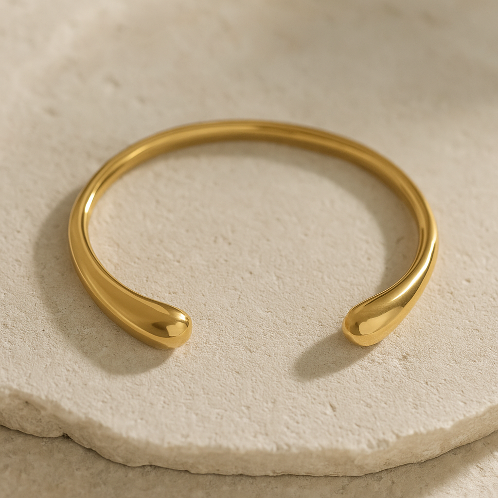 MJ Luxe Ava Gold Cuff Bracelet displayed on beige stone surface, minimalist sculptural open bangle with polished golden finish.