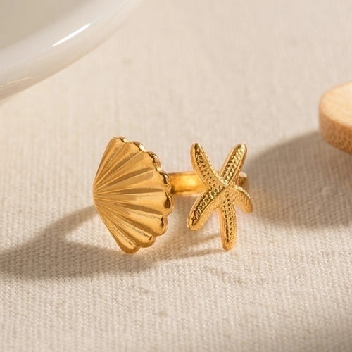 Close-up of the Isla Starfish Seashell Ring, featuring a textured gold starfish paired with a scallop seashell on a neutral fabric background. This adjustable open ring is crafted from 18K gold plated stainless steel, waterproof and tarnish resistant, designed as a coastal-inspired jewelry piece for women. Perfect as a beach-themed ring or ocean inspired gift.